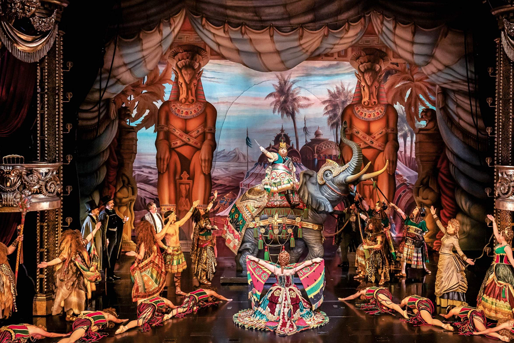 Billet pour le spectacle Le Fantôme de l'Opéra à Londres - Plongez dans le monde enchanteur du Fantôme de l'Opéra au théâtre historique de Londres, le Théâtre de Sa Majesté, où drame et musique se rejoignent pour une expérience live véritablement inoubliable. Dès que le rideau se lève, vous serez entraîné dans l'histoire poignante de Christine, une jeune chanteuse à la voix d'or, et du mystérieux Fantôme qui vit sous l'opéra. Son masque sombre et sa tanière secrète ajoutent une atmosphère de mystère, tandis que son amour profond pour Christine donne lieu à des moments de passion, de tension et de peine sur scène. Admirez les décors époustouflants qui transforment le théâtre en grand halls d'opéra, passages secrets et une tanière souterraine éclairée à la bougie. Les costumes somptueux — robes flottantes, smokings élégants et la cape emblématique du Fantôme — donnent vie aux personnages en couleurs vives. Alors que l'orchestre live emplit la salle de la partition envoûtante d'Andrew Lloyd Webber, vous entendrez des chansons célèbres comme La Musique de la Nuit, Tout ce que je te demande et Pense à moi, chacune interprétée avec une émotion puissante par des chanteurs de classe mondiale. Les effets spéciaux ajoutent une magie supplémentaire : des bougies vacillent sur le lustre du Fantôme, des pétales de rose rouges flottent dans l'air et des ombres dansent sur les murs, créant une atmosphère de romance et de suspense. Chaque instant est conçu pour captiver vos sens, des scènes calmes et tendres aux crescendo palpitants qui feront battre votre cœur. Billet pour le spectacle Le Fantôme de l'Opéra à Londres - Image 5 sur 5